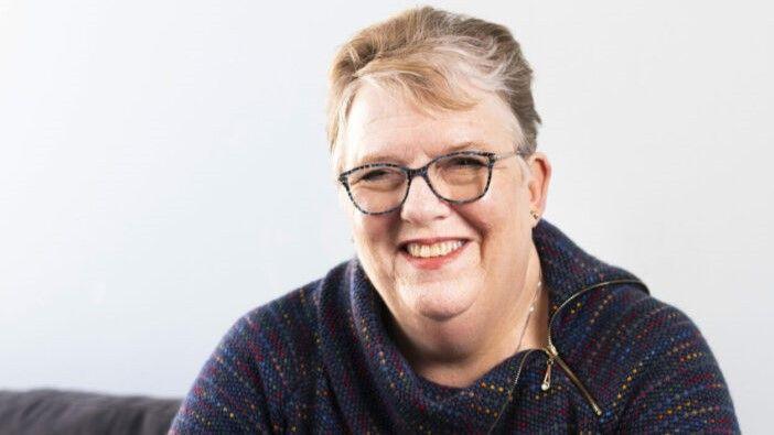 Head and shoulders shot of Susan Wilson sitting down smiling, looking just past the camera. She wears a navy blue knitted jumper with some colourful dots. She has short grey-blonde hair and wears glasses.