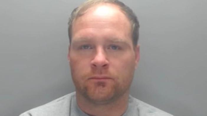 Mugshot of Johnson. He has faint light stubble and a receding hairline with blond hair swept across the top of his head.