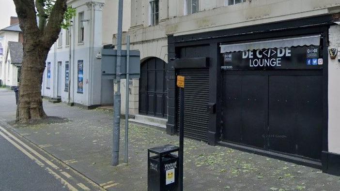 A street with several buildings on it. One of them has black shutters and black signage with "DECODE LOUNGE" printed above the entrance.
