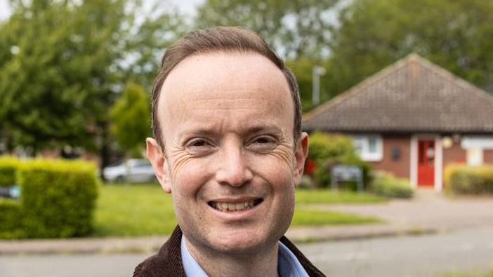 Richard Rout smiles at the camera. He has short brown hair and wears a brown jacket with a blue shirt underneath. He is standing in front of a patch of grass with a bungalow behind him.
