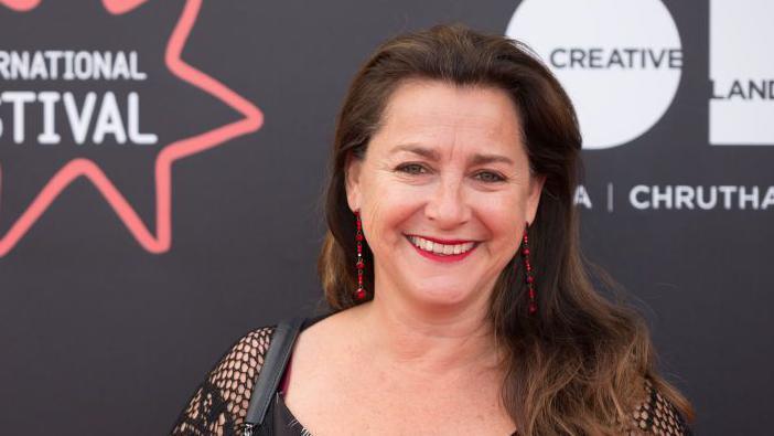 Director Polly Steele wears red lipstick and earrings, she is smiling at the camera and standing in front of a black wall that reads 'Edinburgh International Film Festival.'