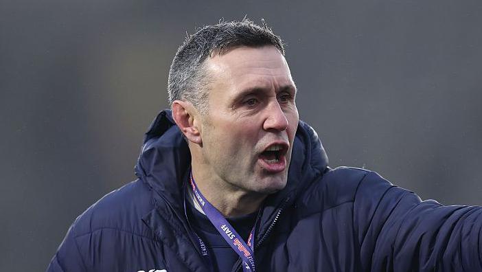 Stephen Jones pointing with his left hand towards players out of shot, while wearing a lanyard and a thick padded coat
