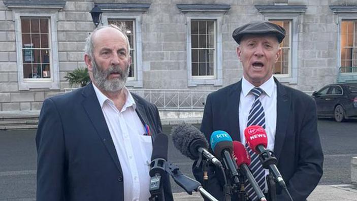 Brothers Danny and Michael Healy-Rae standing in front of a row of microphones. Danny is on the left - he has grey hair and a beard. He's wearing a white shirt and a blazer. Michael is wearing a suit, with a striped tie, and is wearing a flat cap