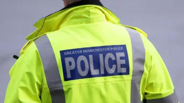 An officer wears a yellow high-vis jacket with the words Greater Manchester Police on the back.