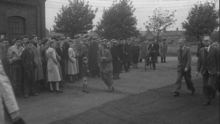 Dozens of people - men and women - stood at the side of the pit entrance waiting for news of their loved ones. The image is in black and white.