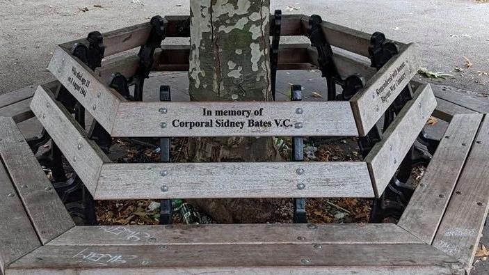 Wooden park bench incribed with words "In memory of Corporal Sidney Bates VC"