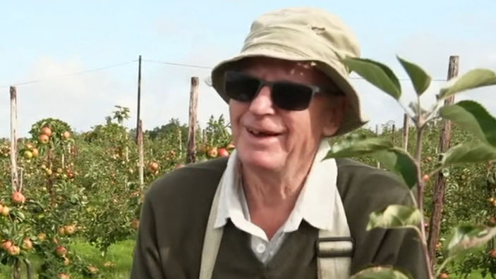Roderick Davis, 79, He is standing in an apple field. He is wearing sunglasses, a hat, and smiling. 