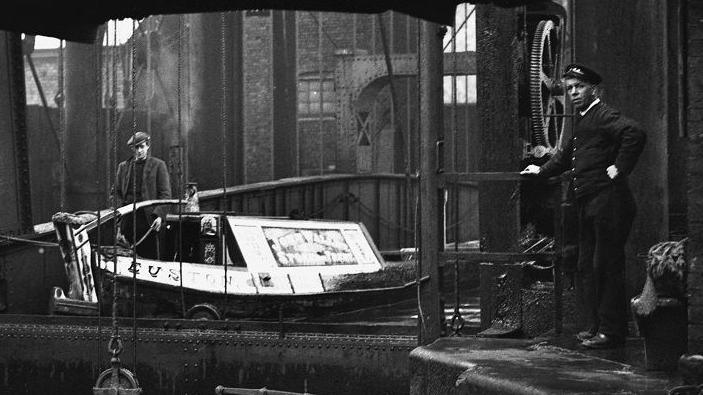 Black and white image showing two men flanking a boat that is being lifted up the iron structure.