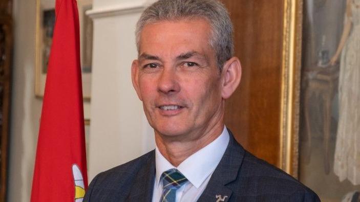Tim Crookall, a man with grey hair and brown eyes, he looks at the camera, there is a white background.
