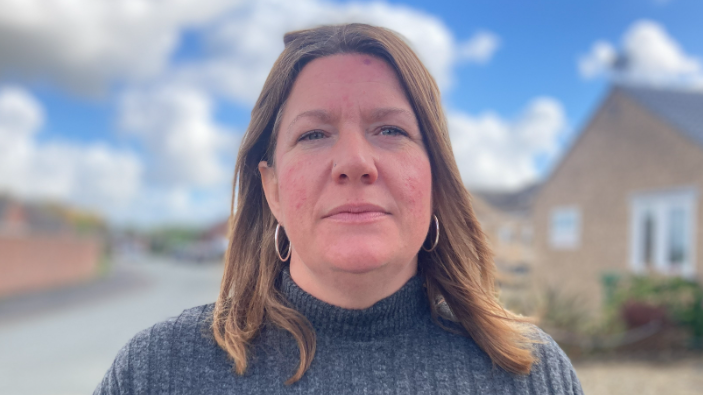 Bryony O'Neill is looking towards the camera. She has shoulder length light brown hair and is wearing gold hoop earrings. She is wearing a grey jumper and standing in front of bungalows. 