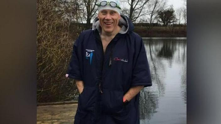 A man wearing goggles, a swim hat and swimming robe is standing on decking in front of a lake