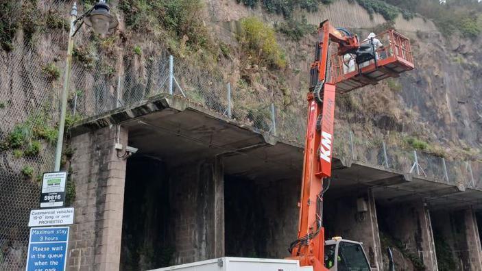 A cliff face is seen with a crane in front of it, on which two people stand in white protective suits in preparation for removing an Asian hornet nest, while in the foreground a lamp post and signage can be seen. 