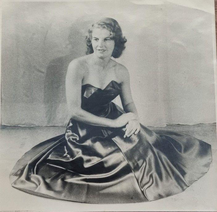 Black and white photo of woman with hair slightly permed and bobbed in a 1940s style. She wears a bustier gown of a dark colour and is sitting on the floor.