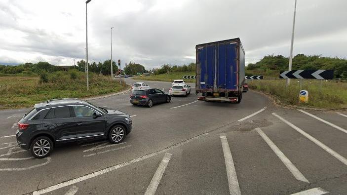 Three-lane carriageway approach to a roundabout. There are white hash lines near the camera. There are cars in all three lanes heading onto the roundabout and a blue lorry is about to enter the roundabout in Lane 3. There are left-pointing arrows to the right of the picture. The centre of the roundabout contains grass, trees and bushes.