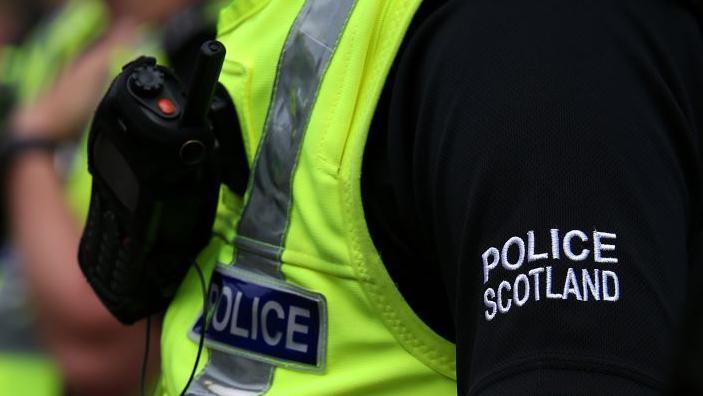 A police officer in a Police Scotland uniform, with the words POLICE SCOTLAND in bold on the sleeve.