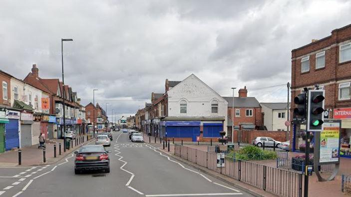 Street view of Bearwood Road with shops on both sides of the road and a number of cars on the road. There is also a pedestrian crossing in the photo, with one traffic light showing with a green light. 