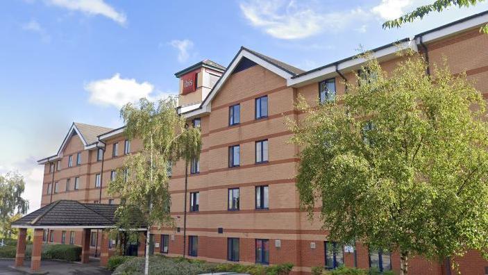 Red brick building of four storeys with pitched roof, with some trees outside. Ibis logo on small tower at the top, under blue sky.