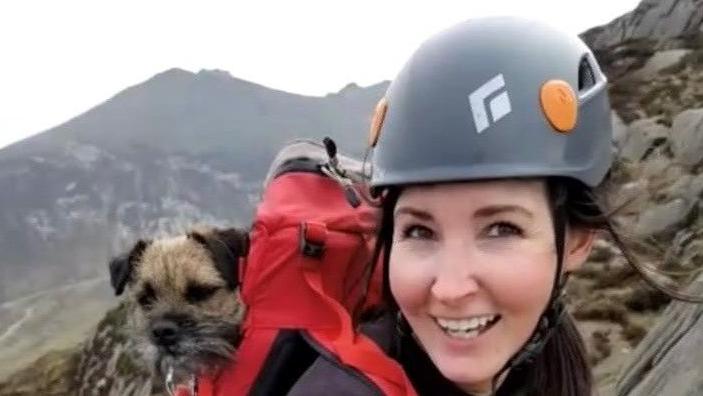 Aly Coyle wearing a helmet, she had long brown hair tied up, she's carrying a red backpack with her dog inside (a terrier type dog with light brown and grey hair). Aly is climbing Cavehill smiling at the camera. 