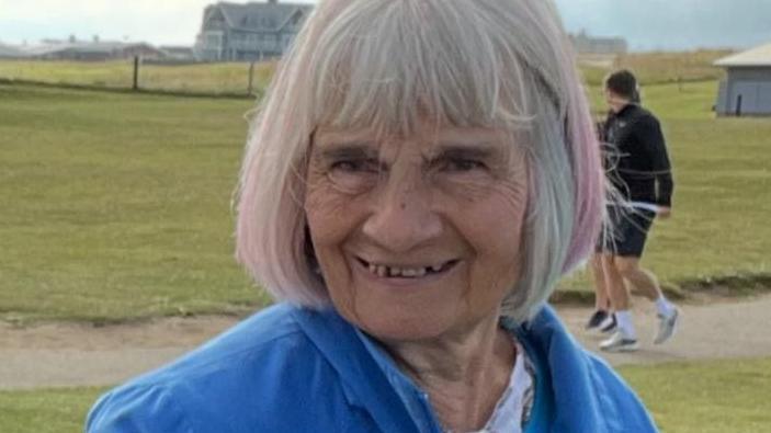 Geraldine Gibson smiles at the camera in a bright blue jacket. She has a short grey bob and fringe and grass and the sea is visible behind her.