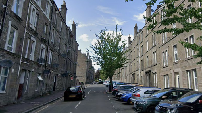 A row of grey tenement buildings with cars parked in bays outside. There are trees dotted at intervals along the road in front of the flats.