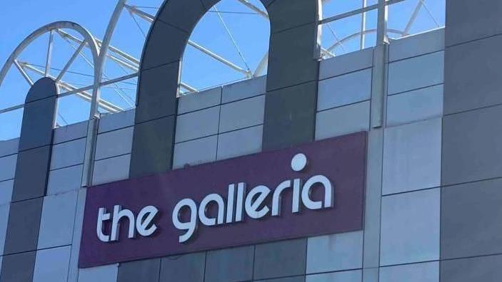 The front of a white-and-grey building with a purple rectangular which has the words "the galleria" written in white, lower-case, letters.