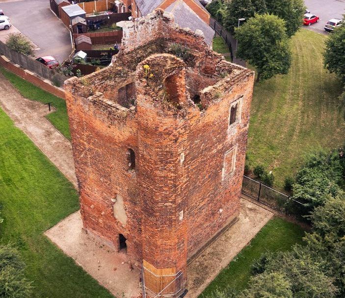 A drone view of Hussey Tower - a red-bricked tower. Its roof is missing. It is surrounded by green grass and trees