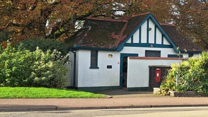 A public toilet is seen, with a mail box in front of it, a road in the foreground and plants and trees around it.