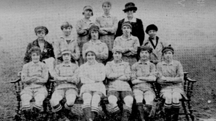 A grainy black and white photo of Helens Ladies AFC pictured in 1921 with the front row sitting on chairs. Other women are standing, some wearing hats and all with their arms folded. 