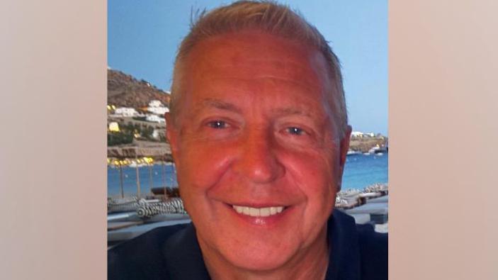 David Barral is smiling at the camera. He has short gery hair and is wearing a dark shirt open at the neck