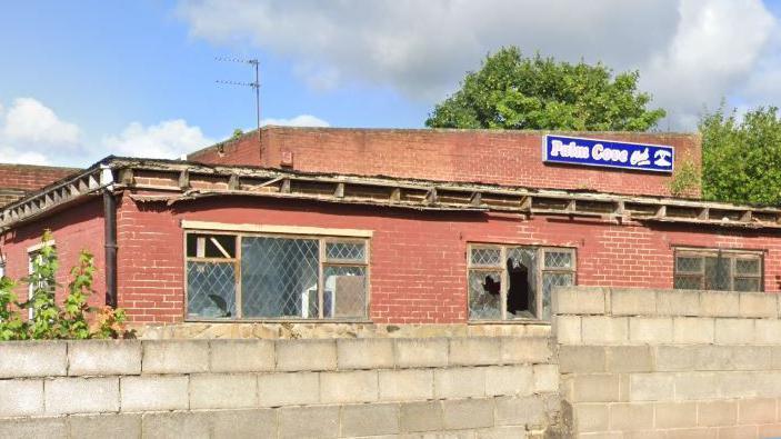 An exterior view of the Palm Cove Club, with a large brick wall in front of the derelict venue. The former club has smashed windows, with the roof partially collapsed. 