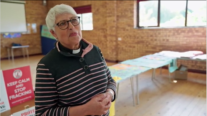 A woman with grey hair and glasses, wearing a dog collar, stands in a hall with a red sign behind her, which reads 'keep calm and stop fracking.' 