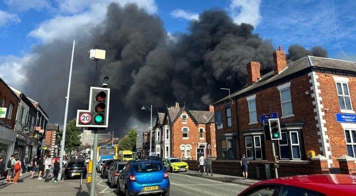 A large plume of black smoke can be seen billowing above rooftops on a street lined with cars as onlookers watch. There is a police car and ambulance on the street too.