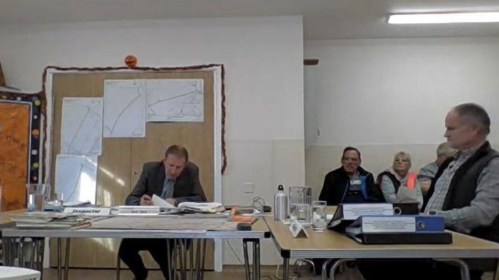 A village hall with modern de-mountable tables arranged for a public inquiry. A man wearing a grey suit sits behind one reading from a book, while other people sit at right angles to him looking at the front. There are maps on the wall pinned up and jugs of water on the tables. 