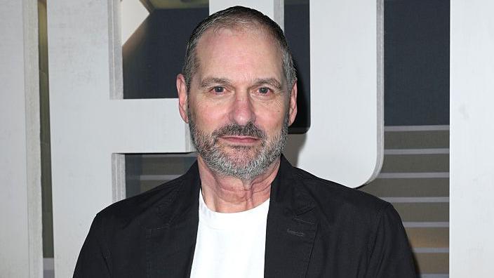 A man wearing a black blazer and white t-shirt stares at the camera. He has short cropped greying hair and a well-trimmed grey beard.