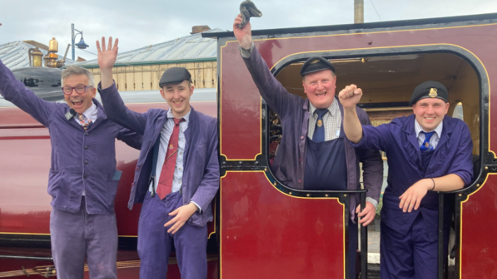 Four men, who are dressed in purple clothing, are standing in a row. Two are on the left and are standing outside of the locomotive, which is painted red and black with gold accents. Two men on the right are standing inside the locomotive.