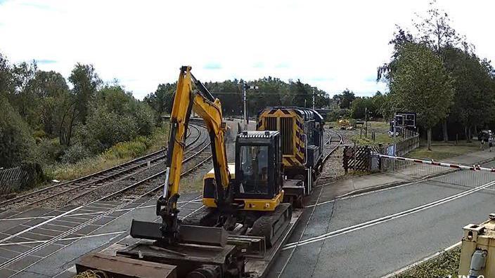 A yellow digger loaded onto a flat, open railway car. It's arm is bent with the bucket resting on a raised platform on the car. A yellow and black engine is visible. The picture is taken at a level crossing and railway tracks, an intersecting road and greenery are visible. 