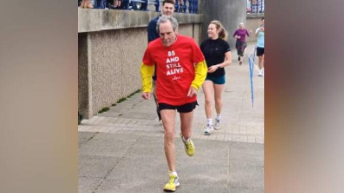 Eric Hughes, wearing a red T-shirt saying "85 and still alive", taking part in the Porthcawl Parkrun