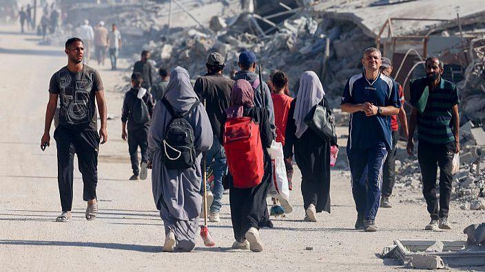Various people walking along in Gaza, mainly away from the camera, with huge piles of debris in the background.