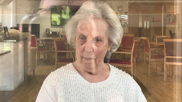 Vivian Harris stares forward while sat in a chair inside a care home. She is wearing a white blouse and has grey hair. The reflection from a window can be seen in the image.