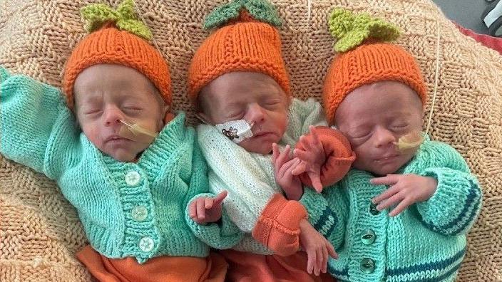 The trio of triplets lie together on a beige-coloured blanket. They are wearing matching green cardigans and knitted orange hats. All three are lying on their backs and have their eyes closed