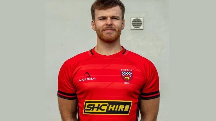 William Bevan. A young man with short hair and a beard wearing a red short-sleeved rugby shirt.