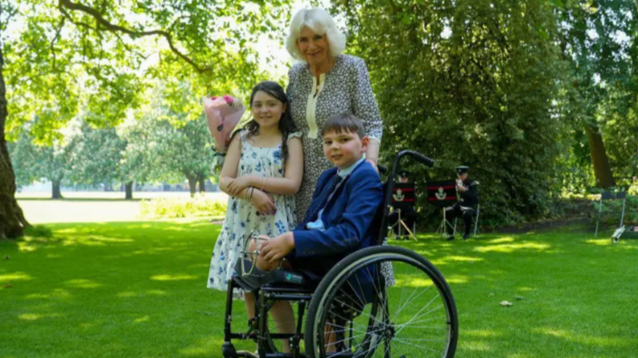 Tony Hudgell with Queen Camilla and Lyla O'Donovan. They have been pictured outside in the sun on the grass. Tony is in a wheel chair - he is an amputee missing both legs. He wears a suit. Lyla has long dark hair and wears a summer dress. Queen Camilla wears a floral dress and is holding a bouquet of flowers. There is a brass band in the background and trees. 