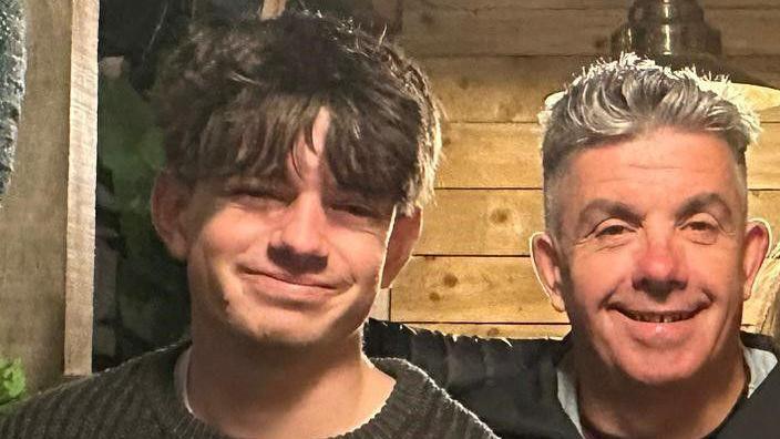 A cropped shot of Sam and his dad smiling at the camera. Sam is wearing a crew-neck knitted jumper and has medium-length brown hair which comes down over his eyebrows. David is wearing a dark coat and has spiky grey hair.