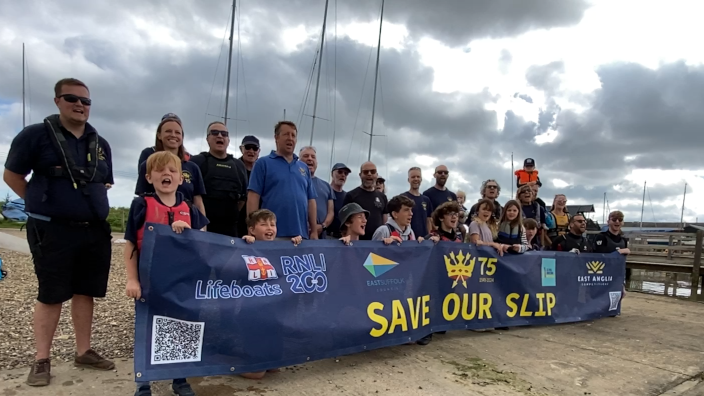 A crowd of people stand under grey leaden skies and behind a blue banner which reads "Save Our Slip".