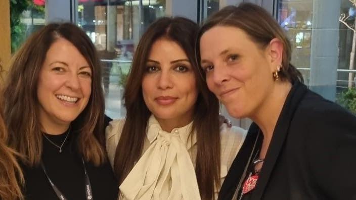 Picture shows three women - Emma, Nour and Jess Phillips. Emma has brown shoulder length hair and is smiling, she wears a black top. Nour is in the middle in a white blouse with a big bow, she has long brown hair and is slightly smiling and Jess Phillips is next to her, also smiling. She wears a dark suit jacket and gold hoop earrings and has short brown hair. 