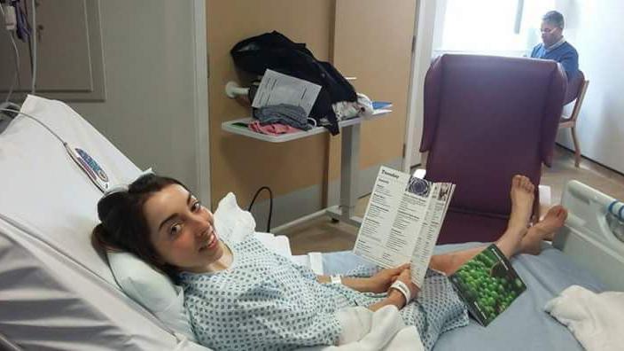 Sophie Wright in a hospital bed, wearing a hospital gown. She is holding an A-level text book and smiling at the camera.