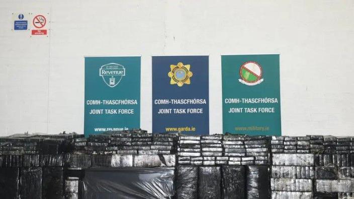 Piles of packages wrapped in black plastic in front of three signs saying 'joint task force'