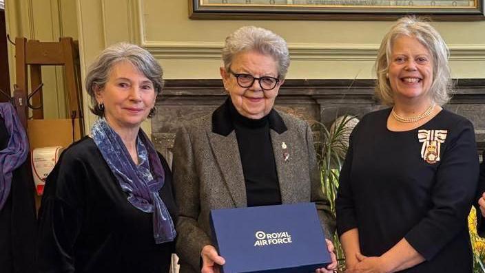 Three women pose for a photo, one in the middle is holding a closed presentation box which reads 'Royal Air Force' on the front.