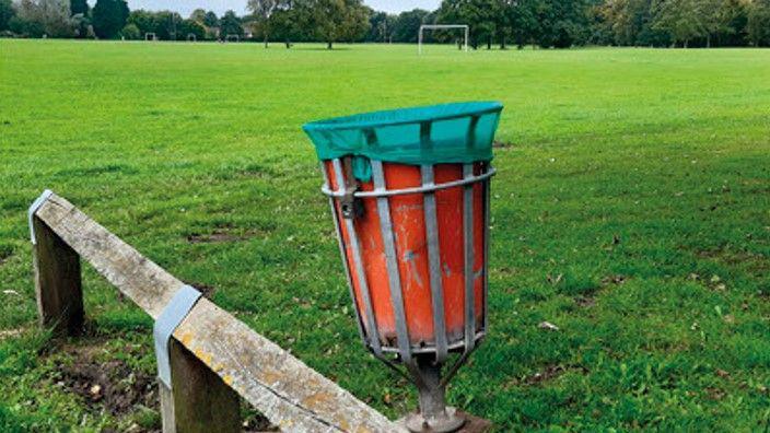 A red bin, which is slightly tipped to one side, is next to a low-level fence with an open space in the background with goal posts and trees in the background.