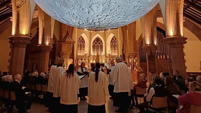 A close-up of the bottom half of the suspended moon, which has visible craters, with the arches of the cathedral on either side and choristers underneath.